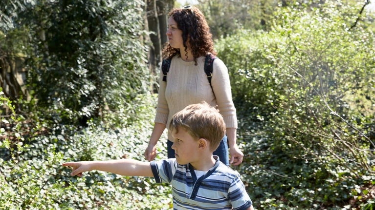 A woman and boy explore a pathway in the sunny garden at Max Gate. The boy is pointing at something through the hedge.